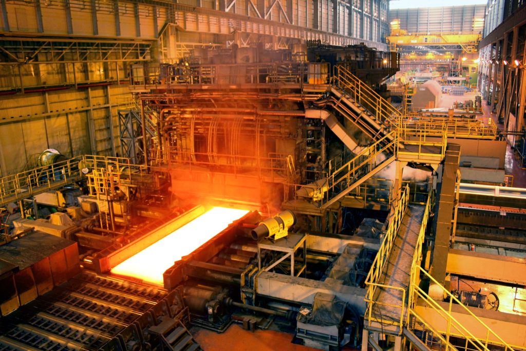 Molten metal glows on a conveyor in a large industrial factory with complex machinery and walkways.