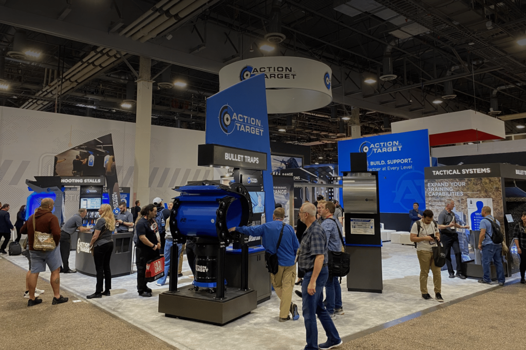 Booth with Action Target branding at trade show, people observing exhibits, indoor convention center.